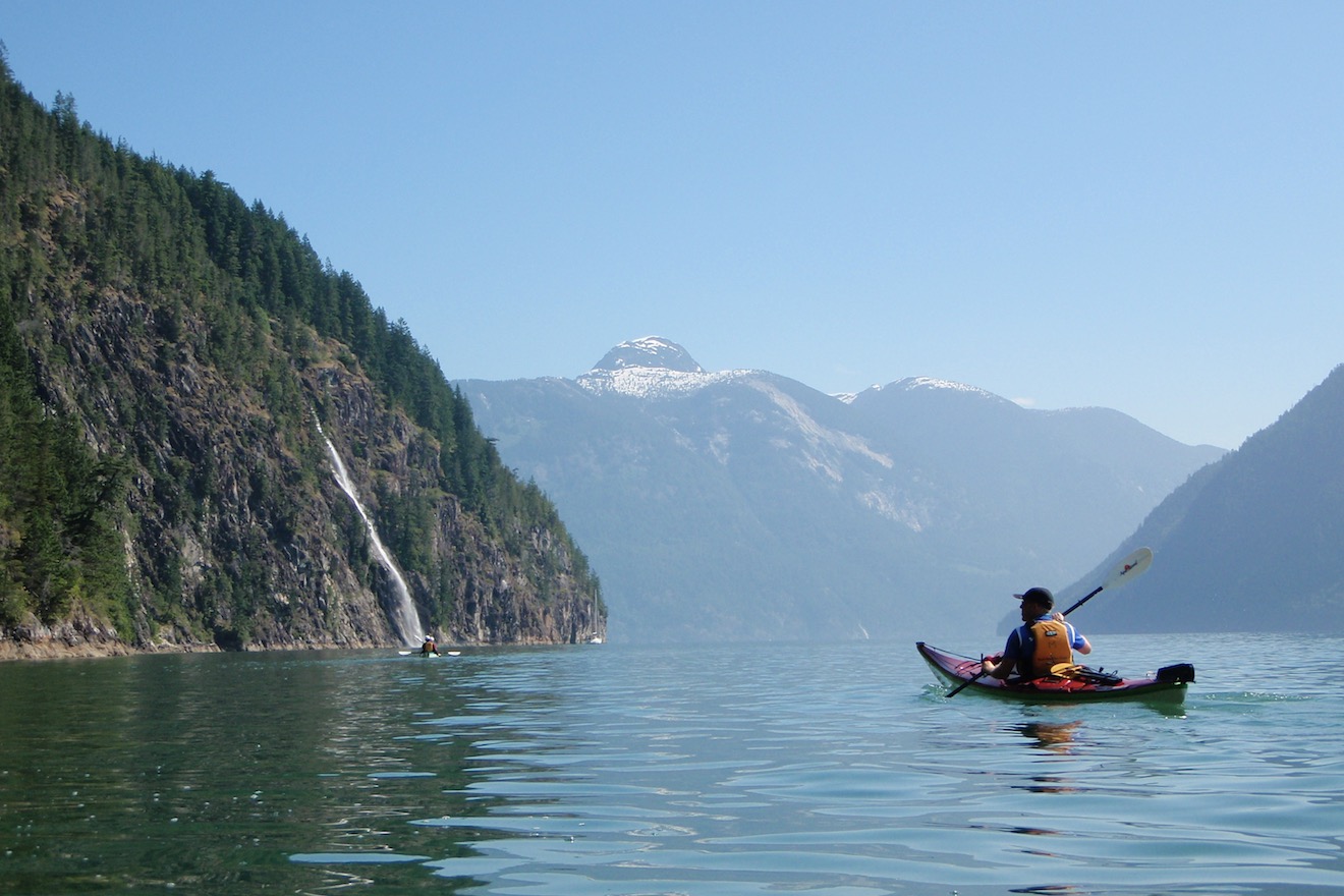 A kayaker paddles towards a large waterfall on a sunny day in Toba Inlet